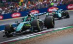 Green Formula 1 car leads a race on a curving track, with a cheering crowd in the stands behind a blue barrier and another car close behind.