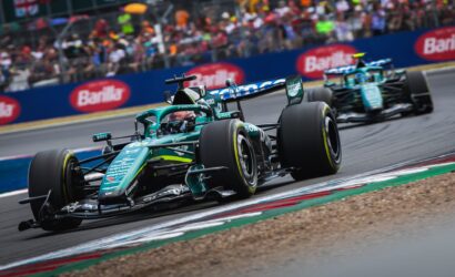 Green Formula 1 car leads a race on a curving track, with a cheering crowd in the stands behind a blue barrier and another car close behind.