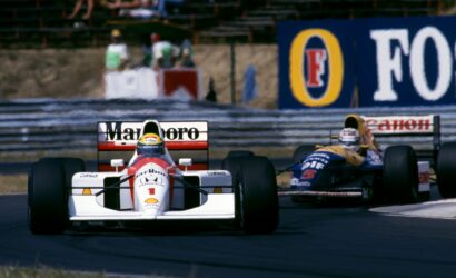 Front-on view of two Formula 1 cars racing on a track, white car with red accents in the foreground and a blue-yellow car behind; sponsor banners along the wall in the background.