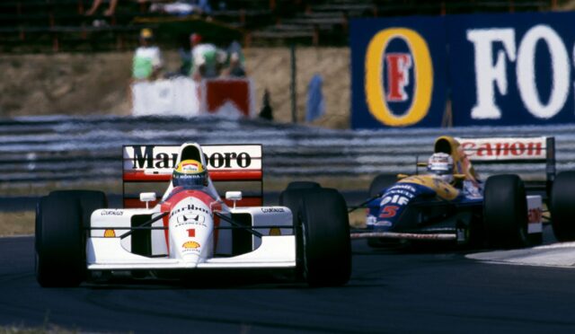 Front-on view of two Formula 1 cars racing on a track, white car with red accents in the foreground and a blue-yellow car behind; sponsor banners along the wall in the background.