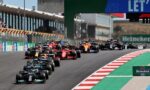 A pack of Formula 1 cars races down a track at the start, red-and-white curbs and green runoff visible, with stands and sponsor boards in the background (Heineken sign).