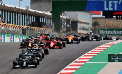 A pack of Formula 1 cars races down a track at the start, red-and-white curbs and green runoff visible, with stands and sponsor boards in the background (Heineken sign).