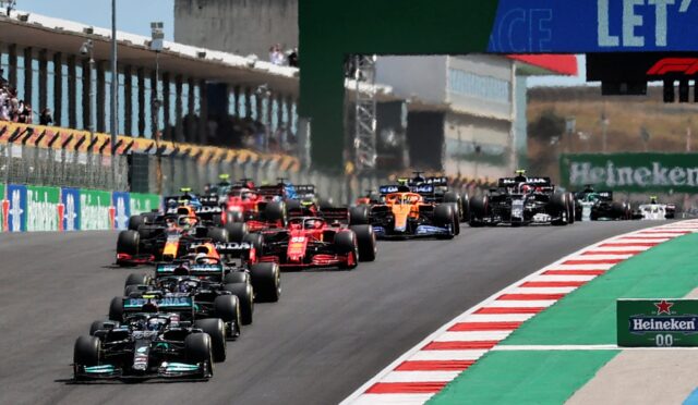 A pack of Formula 1 cars races down a track at the start, red-and-white curbs and green runoff visible, with stands and sponsor boards in the background (Heineken sign).