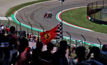 Red Formula 1 car speeds around a curve with cheering fans behind a fence.