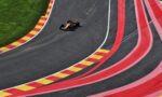 Overhead view of an orange Formula 1 car racing on a curved track with red and yellow striped run-off barriers on the left and green grass beside it.