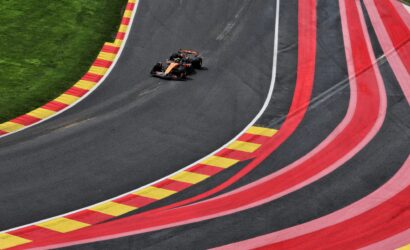 Overhead view of an orange Formula 1 car racing on a curved track with red and yellow striped run-off barriers on the left and green grass beside it.