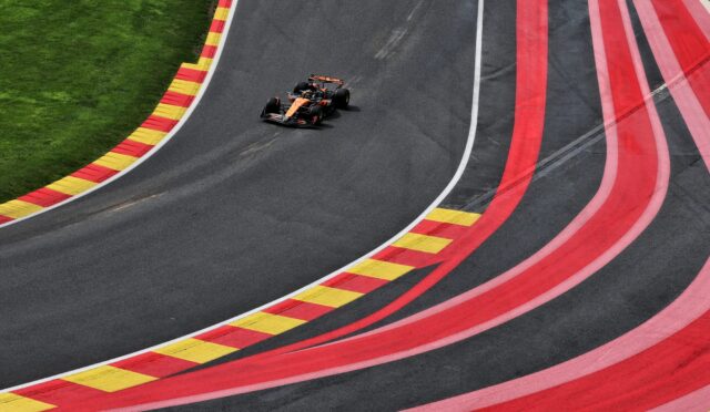 Overhead view of an orange Formula 1 car racing on a curved track with red and yellow striped run-off barriers on the left and green grass beside it.