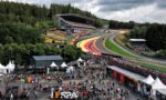 A crowded paddock area at a Formula 1 race with grandstands over the track and colorful corners, surrounded by trees and tents.