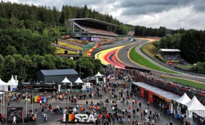 A crowded paddock area at a Formula 1 race with grandstands over the track and colorful corners, surrounded by trees and tents.