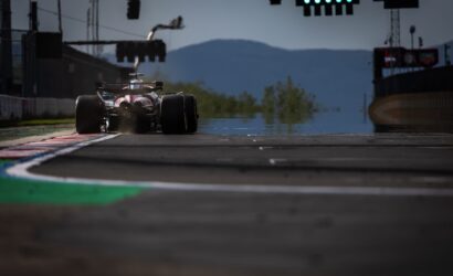 Formula 1 car seen from behind exiting a corner on a race track, smoke behind tires and green circuit lights overhead