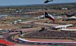 Aerial view of a crowded motorsport event with many cars on a red, white, and blue track and a helicopter overhead.