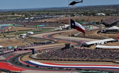 Aerial view of a crowded motorsport event with many cars on a red, white, and blue track and a helicopter overhead.