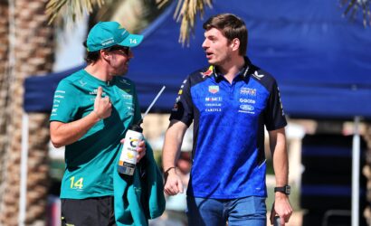 Two men in team jerseys talk outdoors; one in teal with cap holding a water bottle, the other in a blue Red Bull Racing shirt.