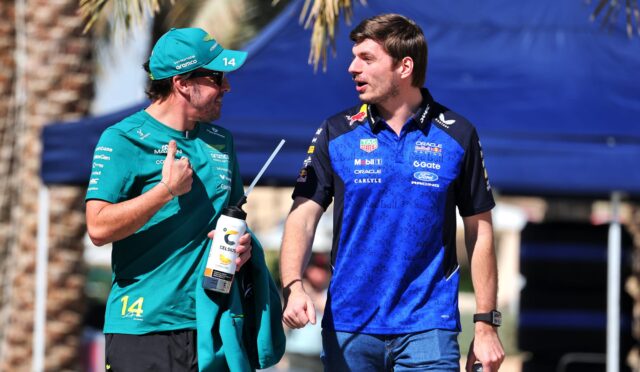Two men in team jerseys talk outdoors; one in teal with cap holding a water bottle, the other in a blue Red Bull Racing shirt.