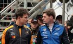 Two race car drivers in orange and blue suits face each other and talk, with a metal staircase behind them.