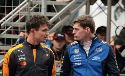 Two race car drivers in orange and blue suits face each other and talk, with a metal staircase behind them.