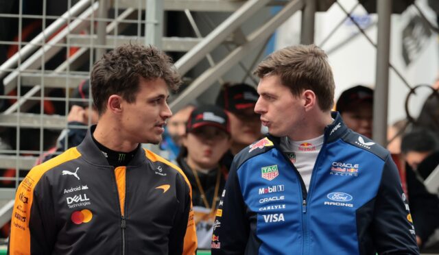 Two race car drivers in orange and blue suits face each other and talk, with a metal staircase behind them.