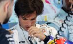Race car driver in a white sponsor jumpsuit wearing a headset, adjusting his gear with crew nearby in a pit area.