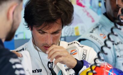 Race car driver in a white sponsor jumpsuit wearing a headset, adjusting his gear with crew nearby in a pit area.