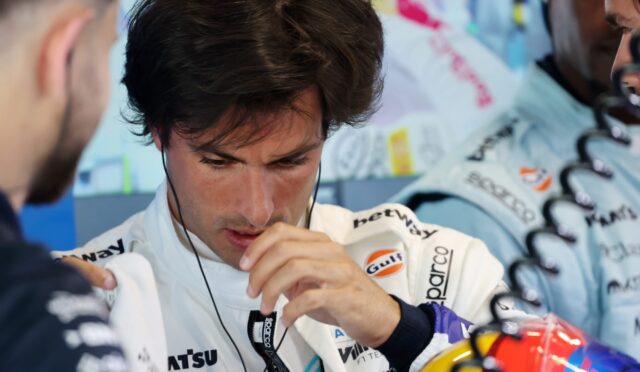 Race car driver in a white sponsor jumpsuit wearing a headset, adjusting his gear with crew nearby in a pit area.