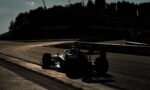 Silhouetted Formula 1 cars on a curving track at dusk; barriers show Lenovo branding and a wooded hill with flags in the background.