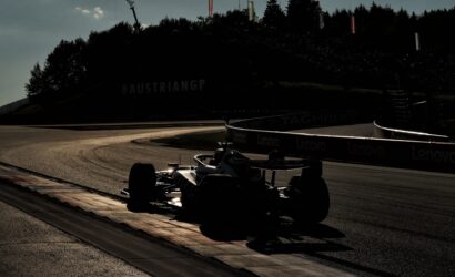 Silhouetted Formula 1 cars on a curving track at dusk; barriers show Lenovo branding and a wooded hill with flags in the background.