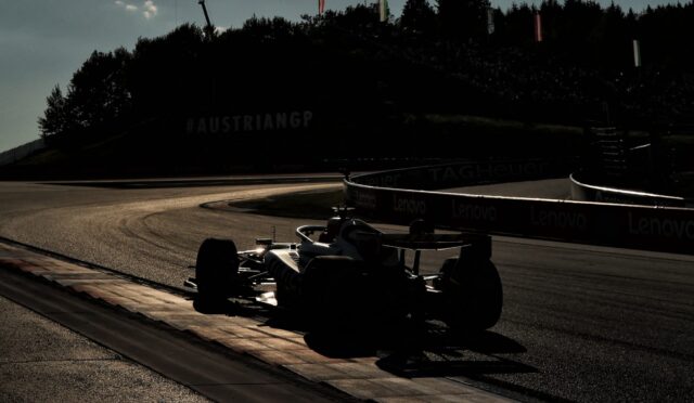 Silhouetted Formula 1 cars on a curving track at dusk; barriers show Lenovo branding and a wooded hill with flags in the background.