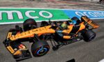 Orange Formula 1 car racing along the track with a colorful green/blue run-off area and spectators in the background.