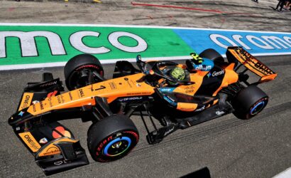 Orange Formula 1 car racing along the track with a colorful green/blue run-off area and spectators in the background.