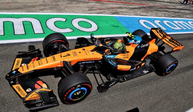 Orange Formula 1 car racing along the track with a colorful green/blue run-off area and spectators in the background.
