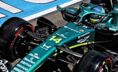 Formula 1 car in teal livery sits at the pit lane with a driver wearing a blue-yellow helmet visible in the cockpit, sponsor logos along the nose and side pods.