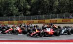 Bruised start of a Formula 1 race with multiple colorful cars skimming the track near yellow Pirelli barriers and a photographer at the edge of the scene.