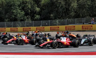 Bruised start of a Formula 1 race with multiple colorful cars skimming the track near yellow Pirelli barriers and a photographer at the edge of the scene.