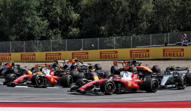 Bruised start of a Formula 1 race with multiple colorful cars skimming the track near yellow Pirelli barriers and a photographer at the edge of the scene.