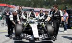 Formula 1 race car being pushed by a team of mechanics in black uniforms on the pit lane, with a crowded grandstand in the background.