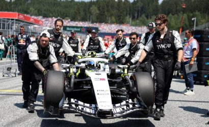 Formula 1 race car being pushed by a team of mechanics in black uniforms on the pit lane, with a crowded grandstand in the background.