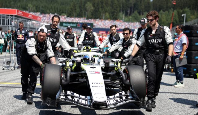 Formula 1 race car being pushed by a team of mechanics in black uniforms on the pit lane, with a crowded grandstand in the background.