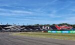 Race track with empty grandstands decorated in black, white, and red, and sponsor banners along the barriers under a clear blue sky.