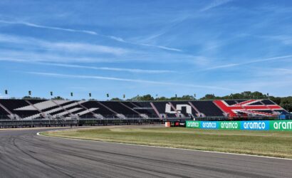 Race track with empty grandstands decorated in black, white, and red, and sponsor banners along the barriers under a clear blue sky.