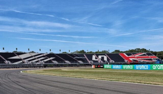 Race track with empty grandstands decorated in black, white, and red, and sponsor banners along the barriers under a clear blue sky.