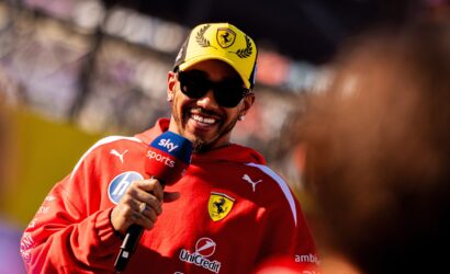 Smiling man in a red Ferrari jersey and yellow cap being interviewed, holding a Sky Sports microphone, at a race event