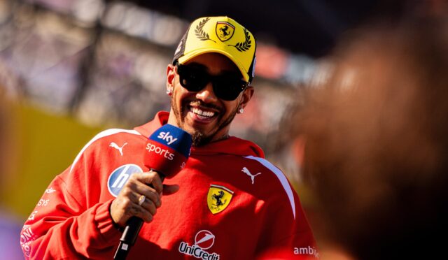Smiling man in a red Ferrari jersey and yellow cap being interviewed, holding a Sky Sports microphone, at a race event