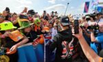 Crowd of racing fans pressed against a barrier, many holding up smartphones to capture the moment; colorful hats and gear abound on a bright day.