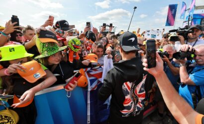 Crowd of racing fans pressed against a barrier, many holding up smartphones to capture the moment; colorful hats and gear abound on a bright day.