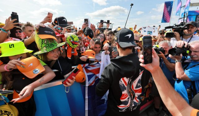 Crowd of racing fans pressed against a barrier, many holding up smartphones to capture the moment; colorful hats and gear abound on a bright day.