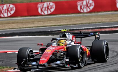 Red Ferrari Formula 1 car speeds around a bend on a race track, with sponsor banners along a red barrier in the background.