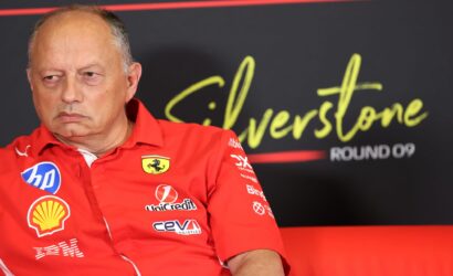 Close-up of an older man in a red Ferrari jacket at a media event, sponsor logos visible on his chest.