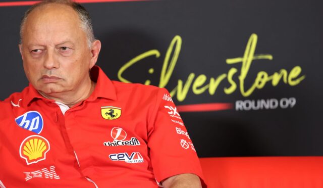 Close-up of an older man in a red Ferrari jacket at a media event, sponsor logos visible on his chest.