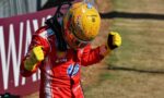 Race car driver in a red suit with yellow gloves celebrates with fists raised near a track barrier.