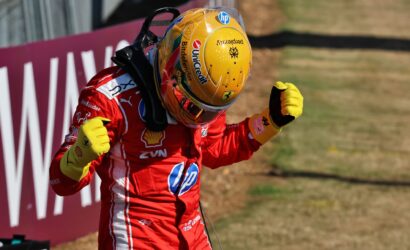 Race car driver in a red suit with yellow gloves celebrates with fists raised near a track barrier.
